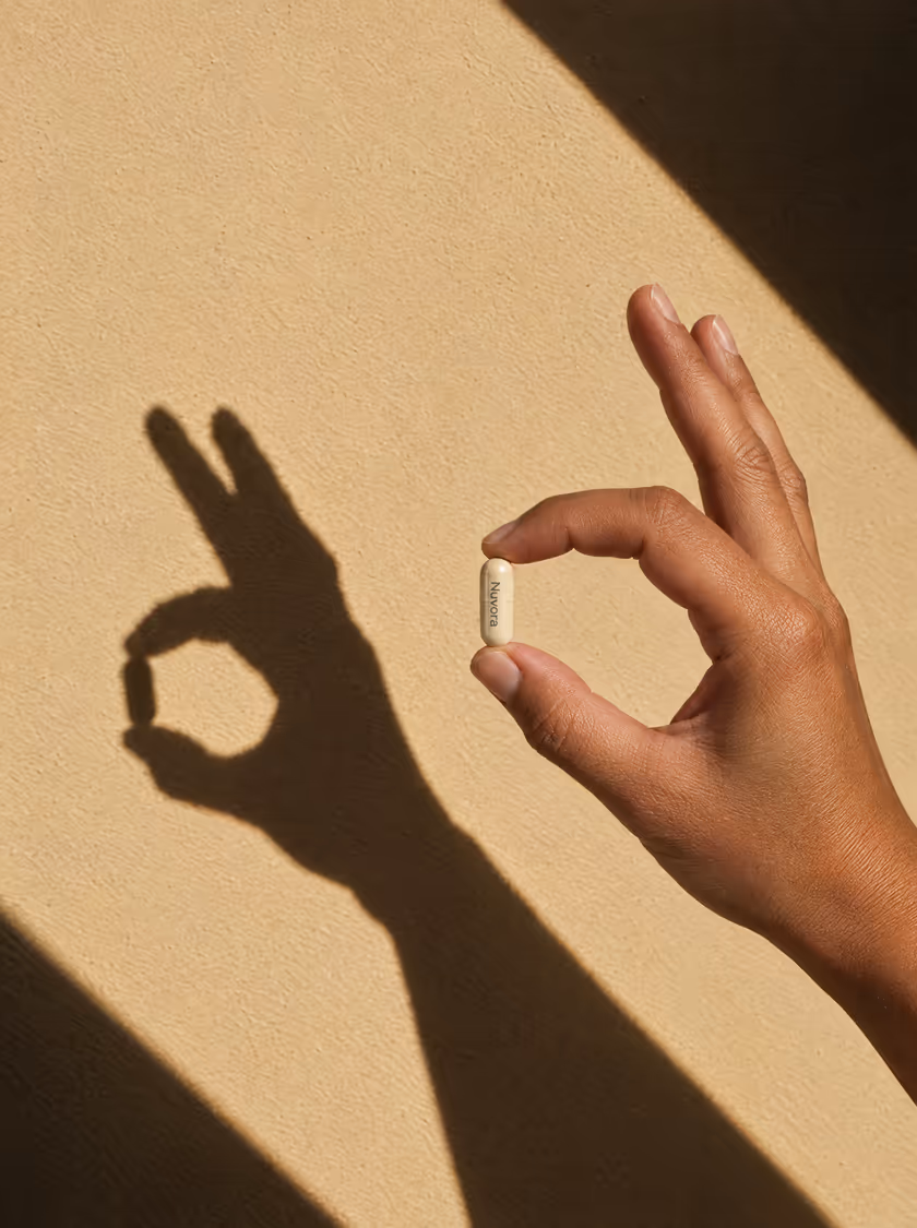 A hand making an OK sign with a Nuvora capsule, its shadow on a sunlit wall